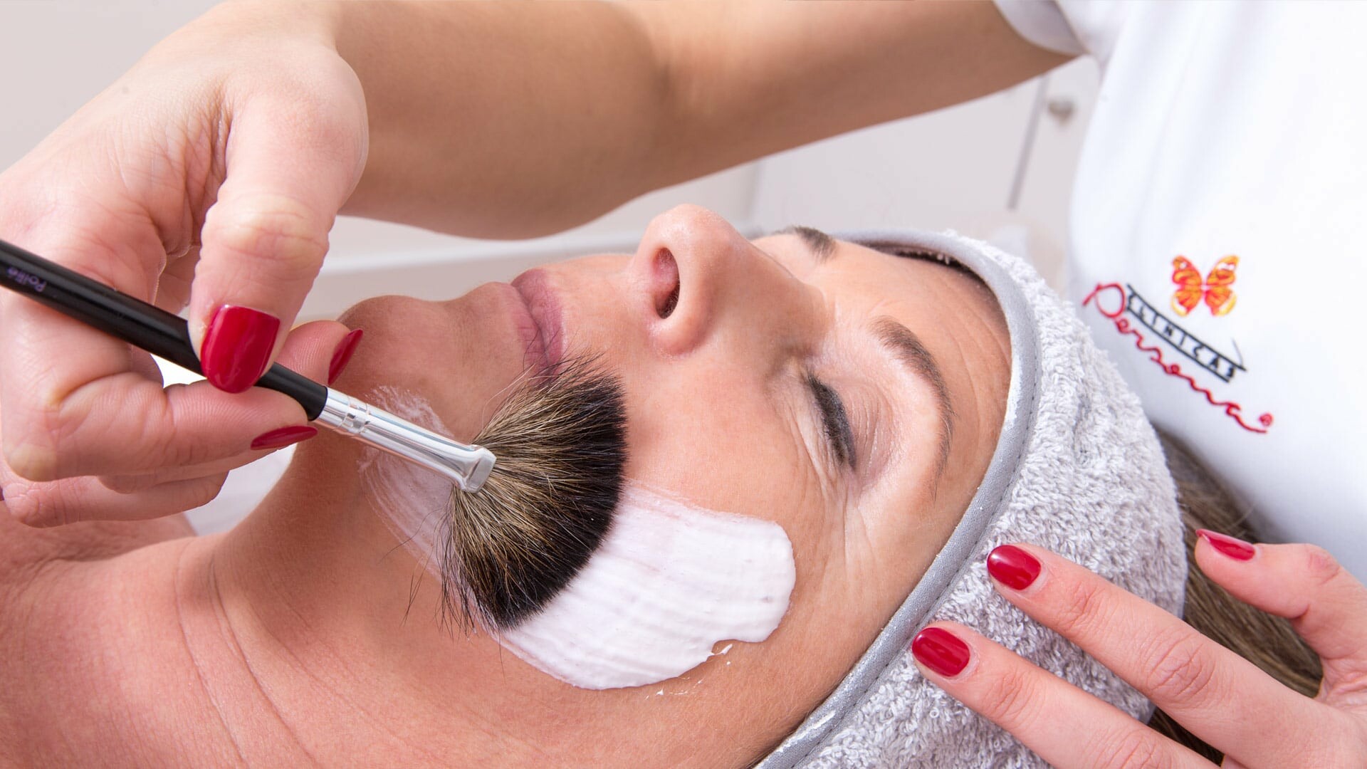 Uma pessoa recebe um tratamento facial enquanto uma esteticista aplica uma máscara branca cremosa na bochecha usando um pincel de maquilhagem. A cliente, usando uma bandolete cinzenta, relaxa de olhos fechados enquanto experimenta a radiofrequência com mesoterapia sem agulhas.