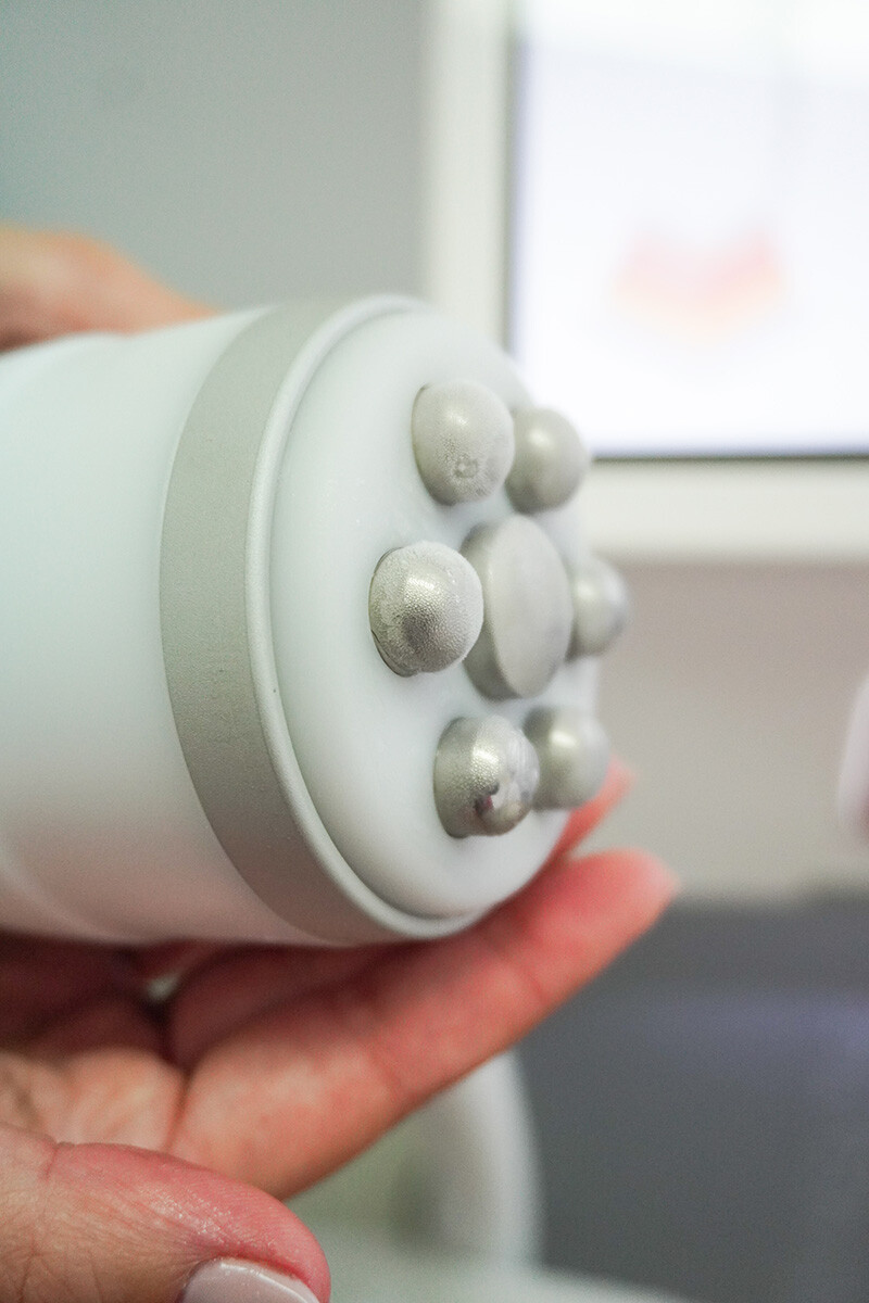 Grande plano de uma mão que segura um dispositivo Pershape® Power com vários nós redondos de metal prateado numa extremidade, possivelmente uma ferramenta de cuidados da pele ou de massagem, contra um fundo desfocado.